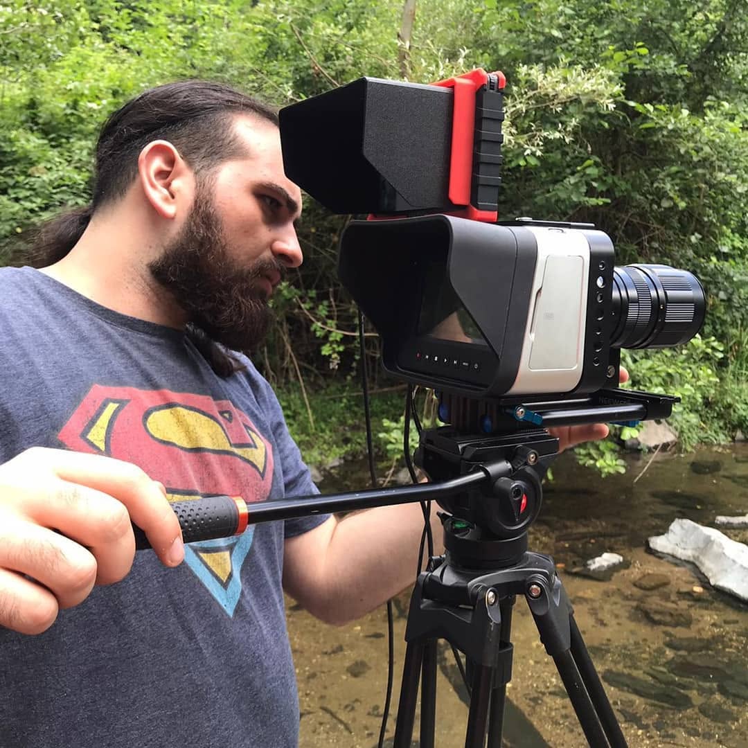 Vittorio Russo, in profile, adjusts a cinema camera on a tripod beside a stream among the vegetation.