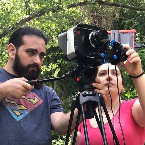 Vittorio Russo holds the handle of a cinema camera on a tripod in the woods, beside another person adjusting the lens.