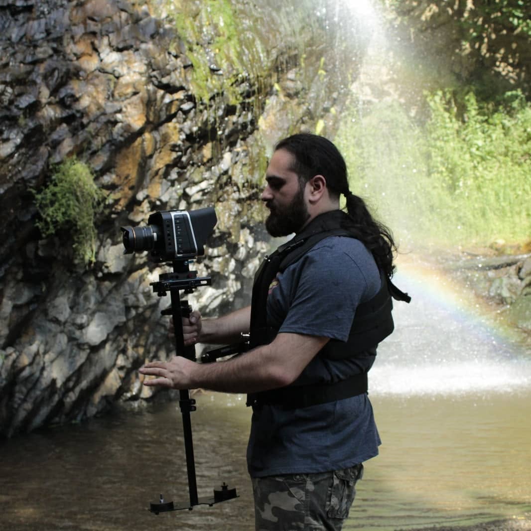 Vittorio Russo, in profile and wearing a vest, holds a stabilizer with a cinema camera in a stream, between a waterfall and a rainbow.