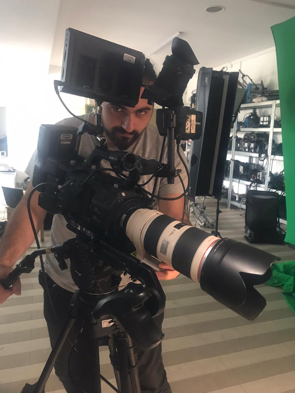 Vittorio Russo stands behind a large cinema camera with a telephoto lens, in a studio with a green screen and equipment shelves.