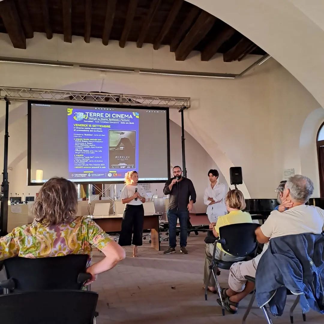 Vittorio Russo speaks into a microphone in a hall with exposed beams, in front of a screen reading "TERRE DI CINEMA".