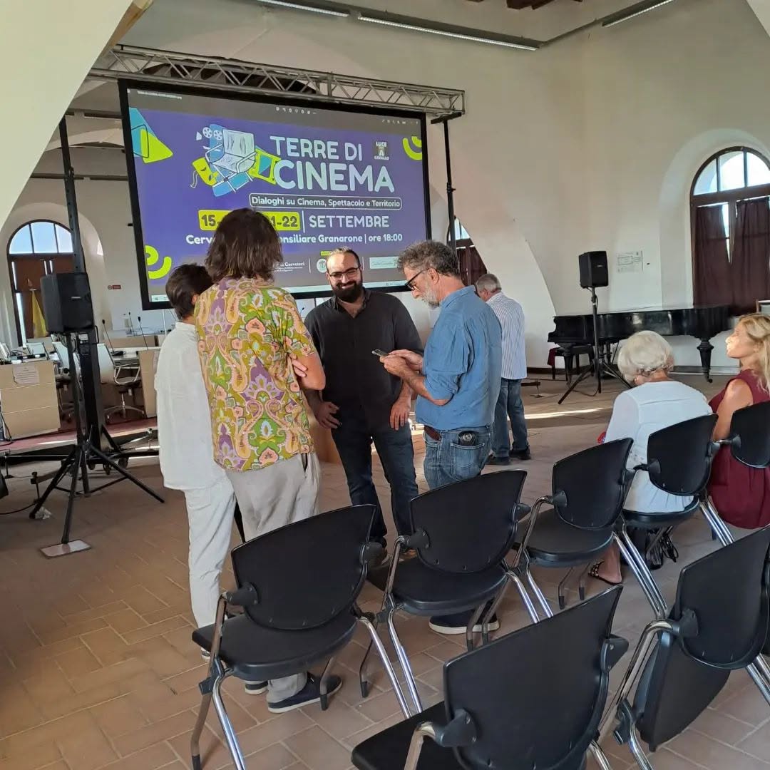 Vittorio Russo, wearing glasses, stands talking with other people in a hall, in front of a screen reading "TERRE DI CINEMA".