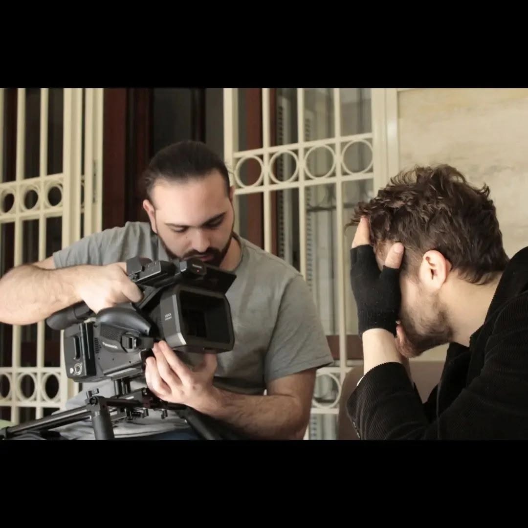 Vittorio Russo adjusts a cinema camera while, beside him, another person holds their head with one hand.