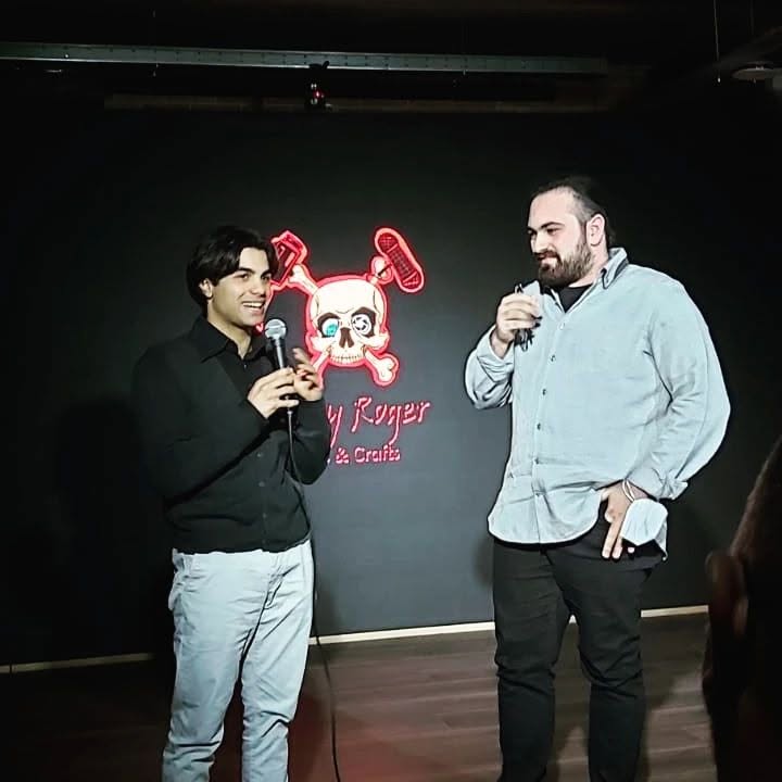 Vittorio Russo holds a microphone on a dark stage, beside another person, in front of a neon sign with a skull.