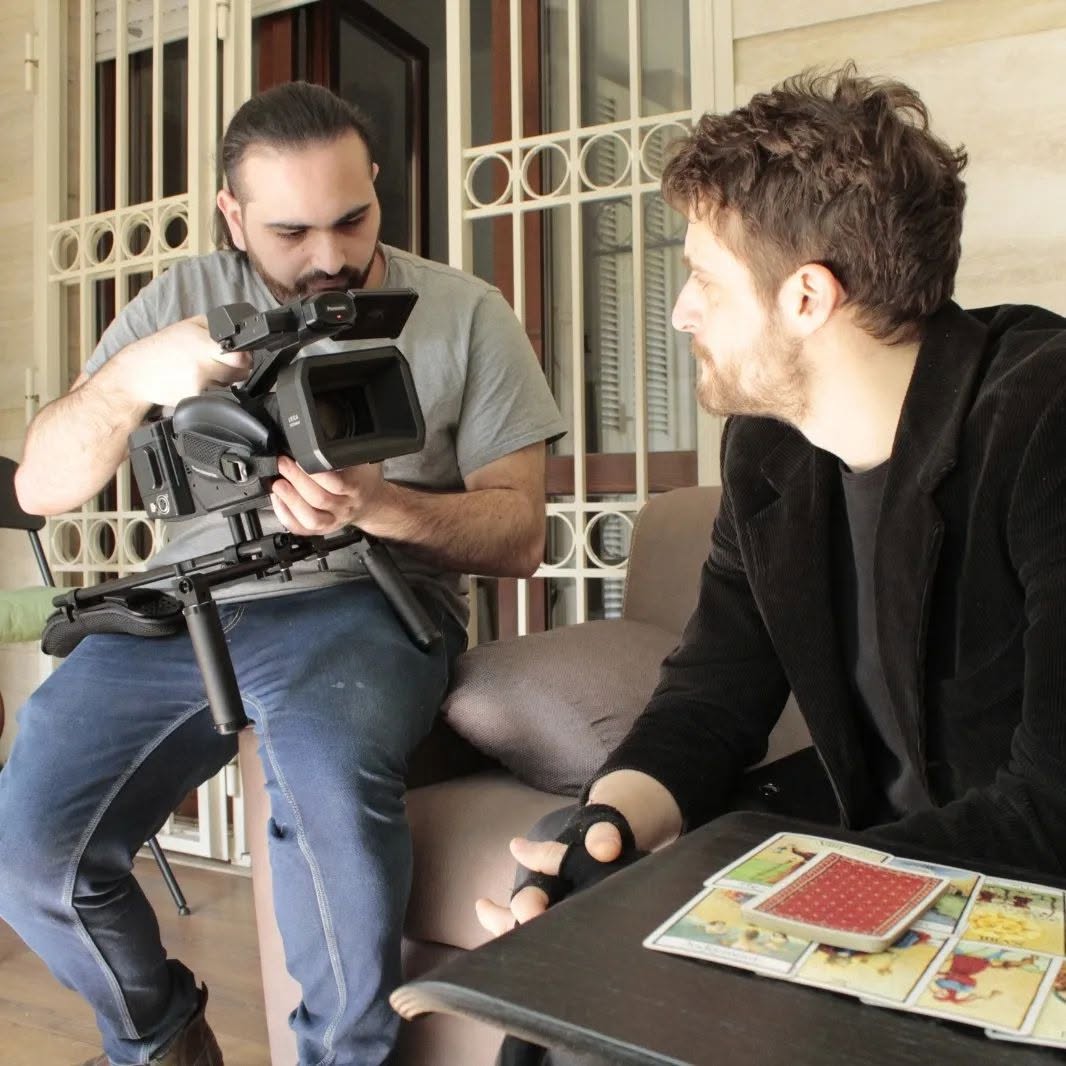 Vittorio Russo holds a cinema camera while seated on a sofa, facing another person, with playing cards on the table.