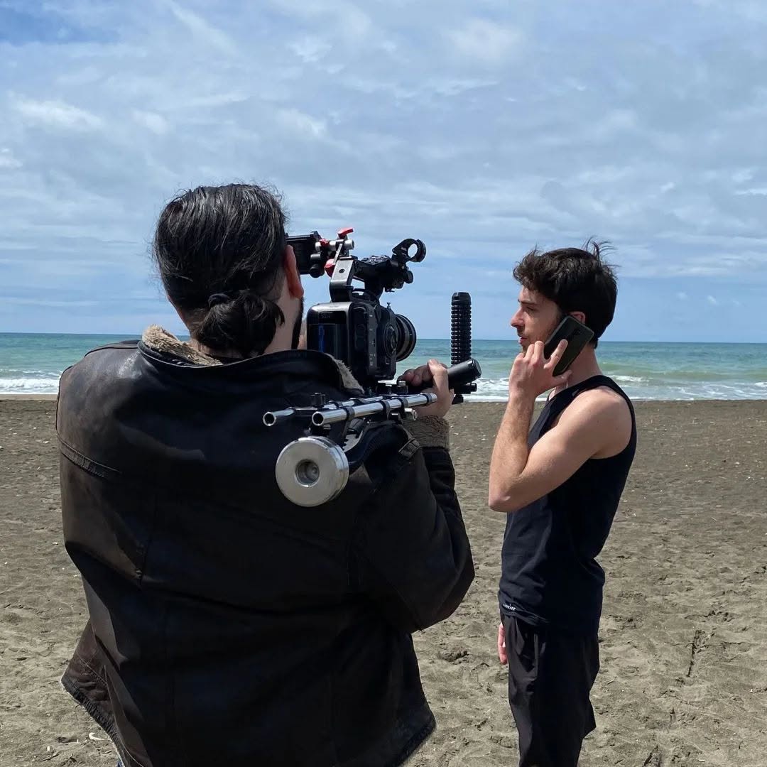 From behind, a person with tied-back hair films a man in a tank top on the beach as he holds a phone to his ear.
