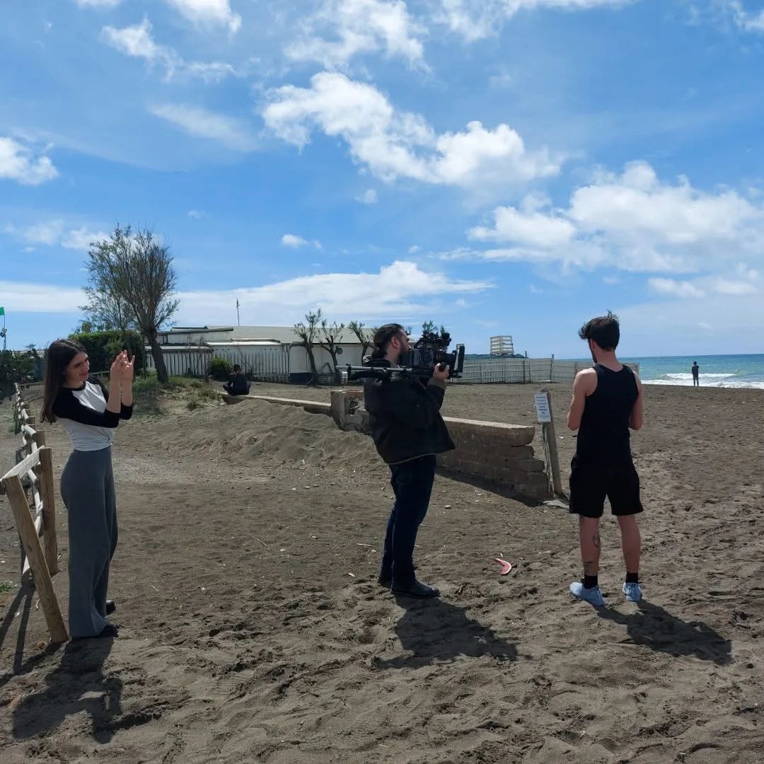 Vittorio Russo films on the beach with a camera on his shoulder, between two standing people, with the sea behind them.