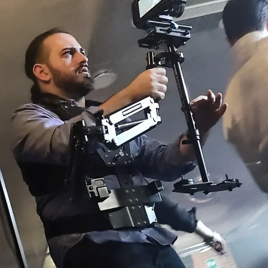 Vittorio Russo, wearing a support vest, holds a stabilizer arm and a cinema camera aimed upward indoors.