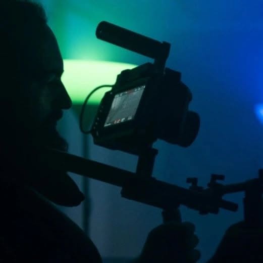Against blue and green backlight, Vittorio Russo in silhouette holds a cinema camera on a rig, its monitor lit.