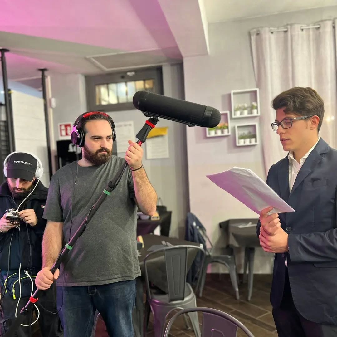 Wearing headphones, Vittorio Russo holds a boom microphone indoors while a colleague films and a man reads a sheet of paper.