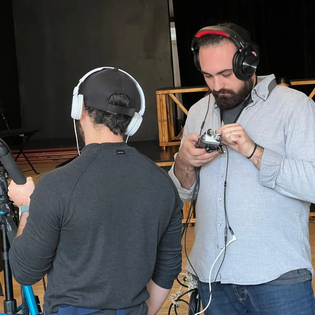 Wearing headphones, Vittorio Russo adjusts an audio recorder beside a colleague seen from behind, in a room with a stage.