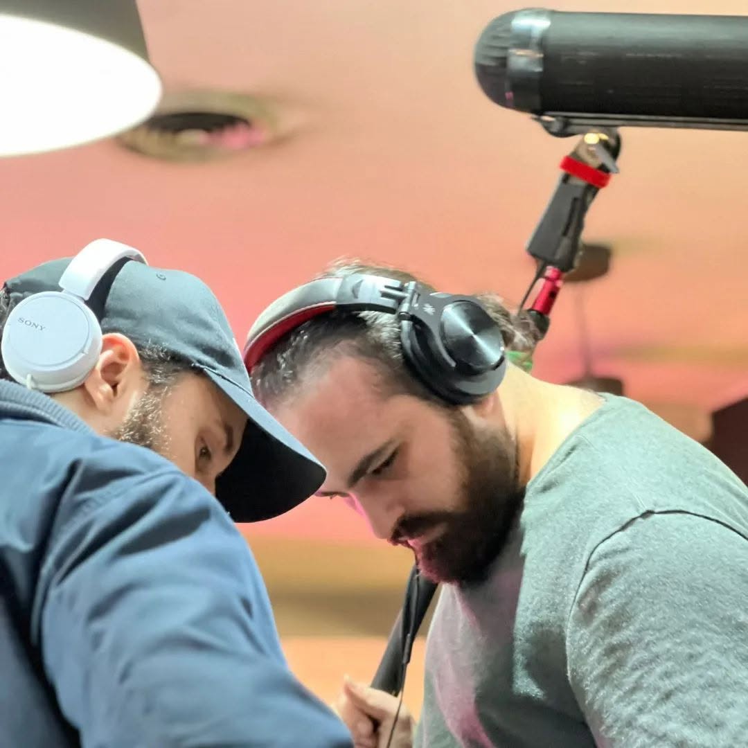 Vittorio Russo, headphones on and hair tied back, leans in with a colleague under a boom microphone in pink light.