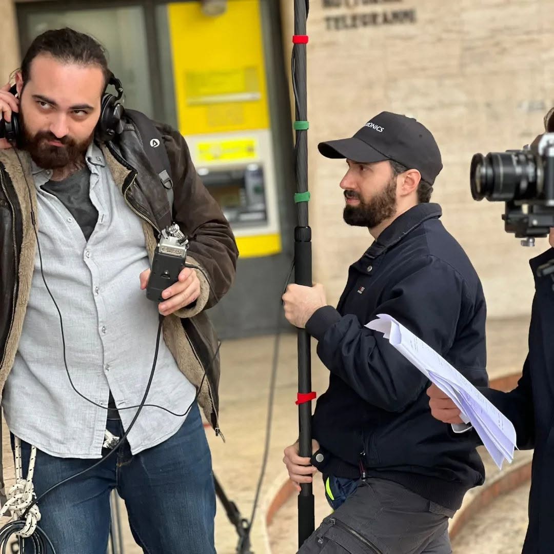 Wearing headphones, Vittorio Russo holds an audio recorder; beside him, a colleague in a cap holds a pole and papers.