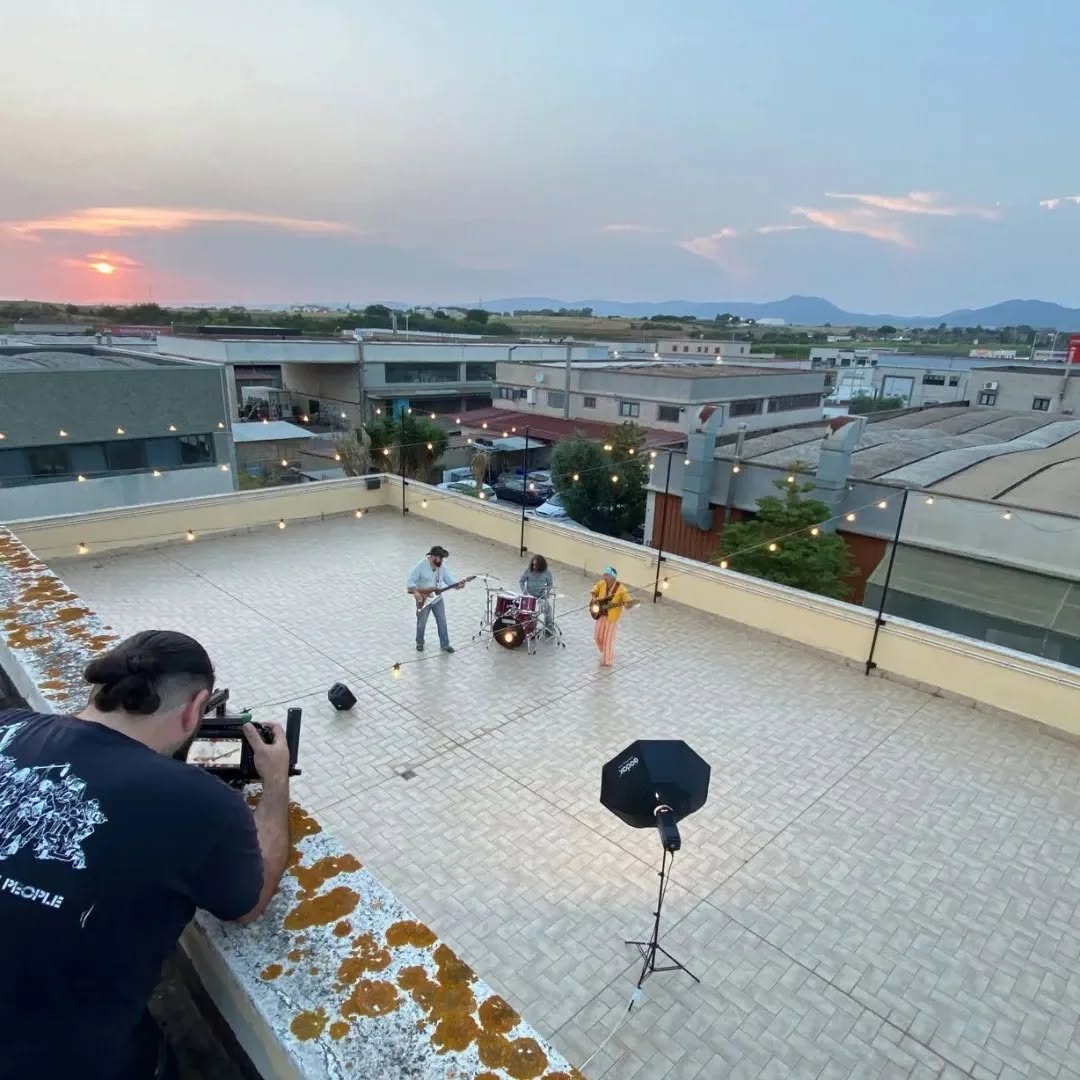 At sunset on a rooftop, Vittorio Russo films a trio with guitars and drums among hanging lights and the rooftops.