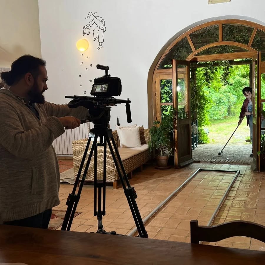 Vittorio Russo adjusts a cinema camera on a tripod in a room with floor tracks, while beyond the arch a person holds a broom.