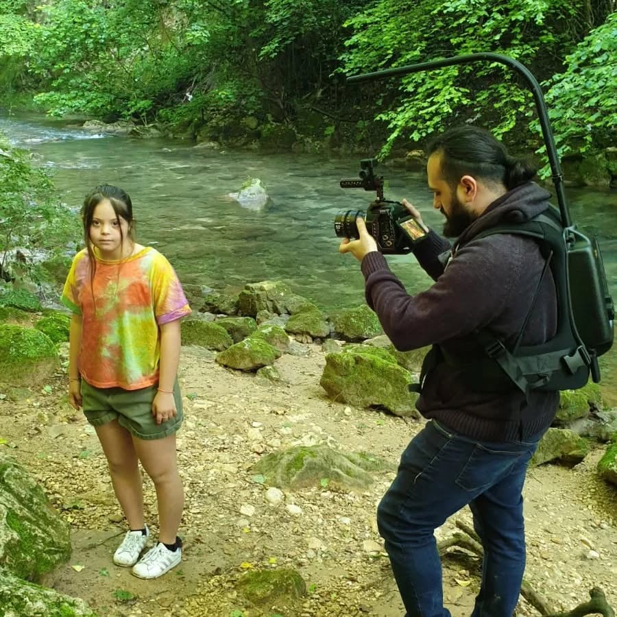 On a stream bank, Vittorio Russo films with a handheld cinema camera a person standing among the rocks.