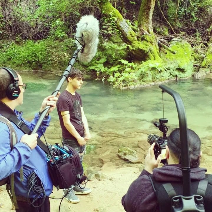 On a stream bed, an operator seen from behind films a standing person while a colleague holds a boom microphone.