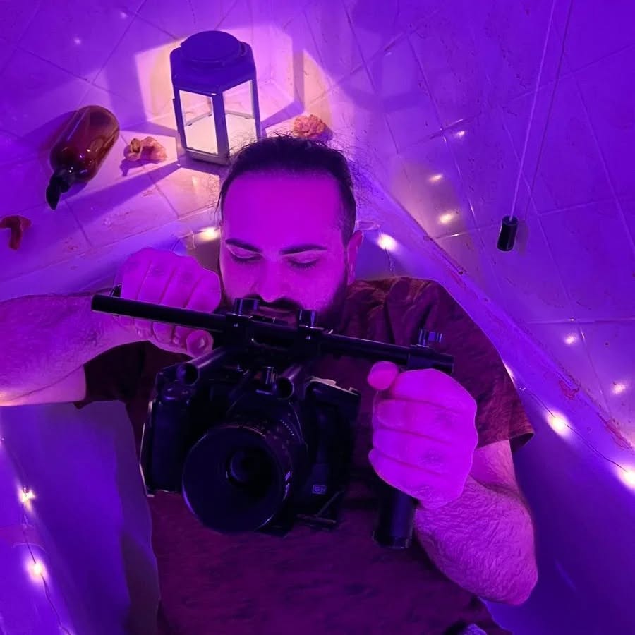 Under purple light, Vittorio Russo lies in a bathtub holding a cinema camera pointed upward, among a lantern and string lights.