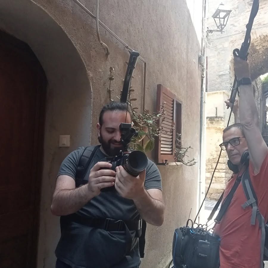 In an alley, Vittorio Russo holds a cinema camera at chest height while a colleague raises a pole.