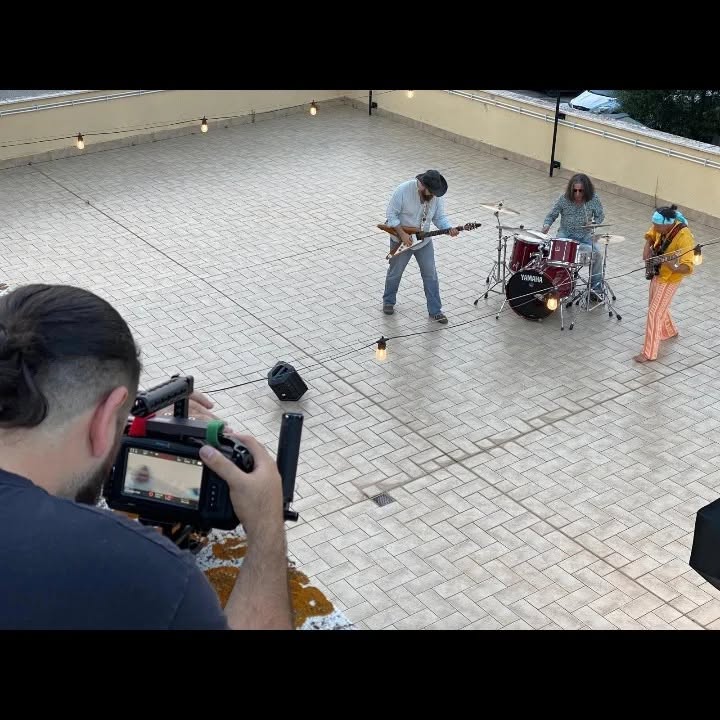 From a terrace, Vittorio Russo films a trio with two guitars and a drum kit, beneath lights hung along the railing.