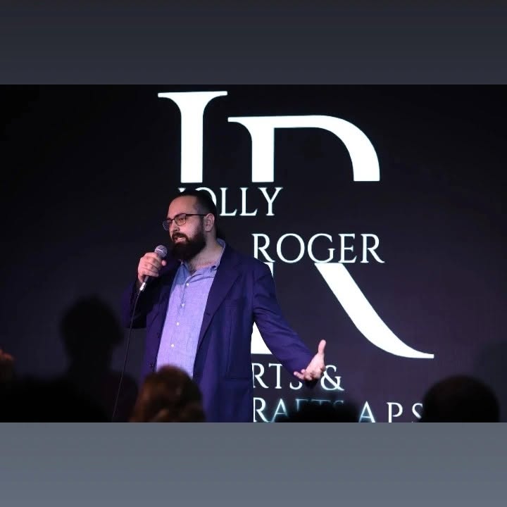Wearing glasses, Vittorio Russo speaks into a microphone on a stage before a screen where “JOLLY ROGER” is readable.