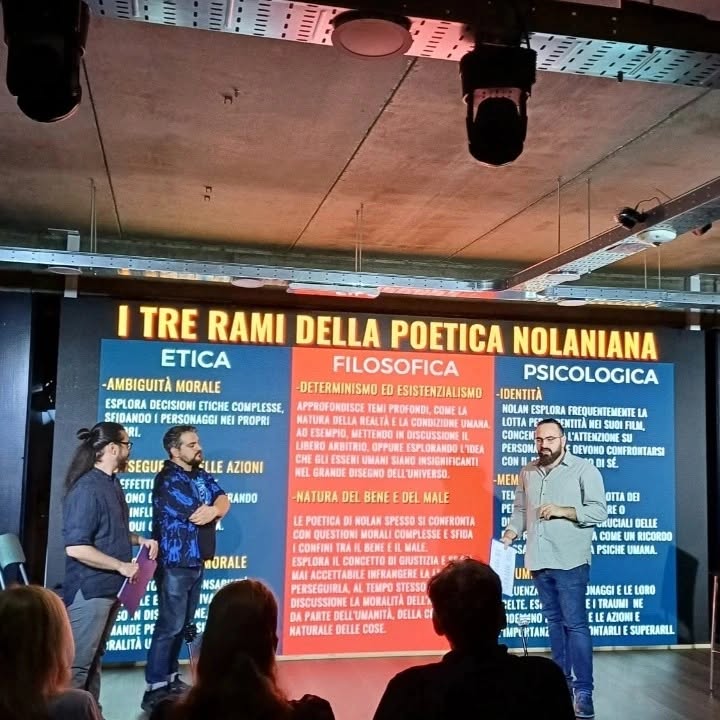 Wearing glasses, Vittorio Russo stands on stage before the screen titled “I TRE RAMI DELLA POETICA NOLANIANA”.