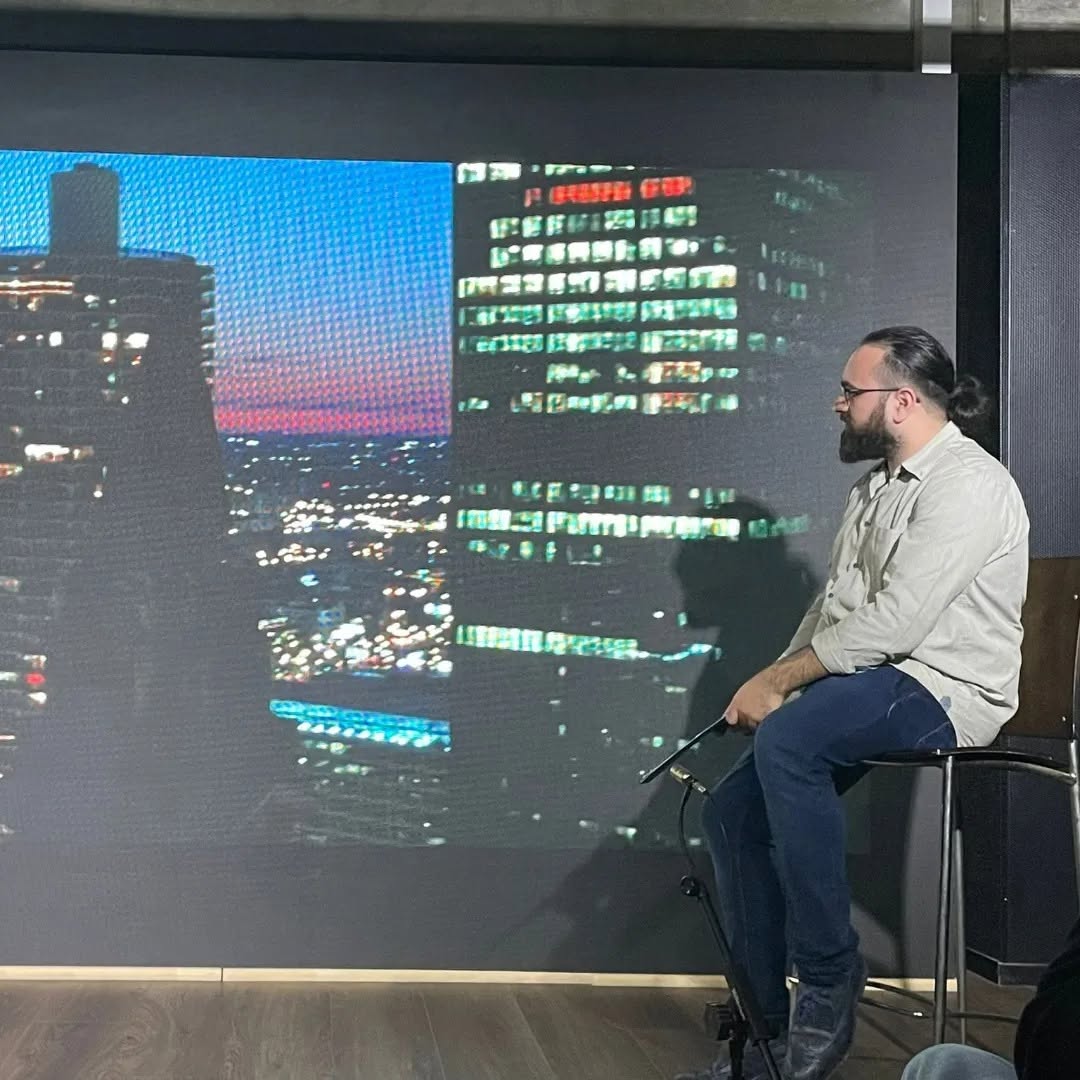 Wearing glasses, Vittorio Russo sits in profile on a stool before a screen showing a city at night.