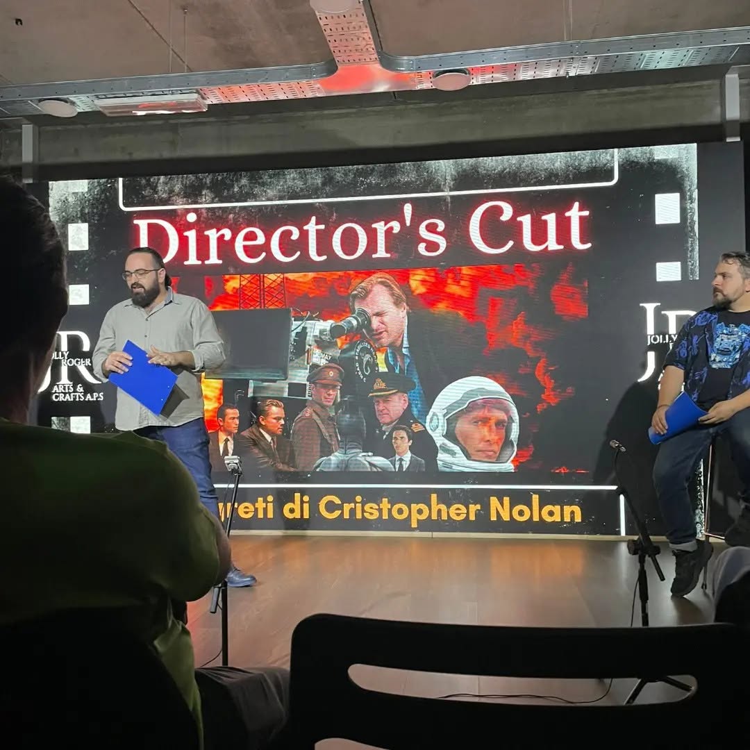 Wearing glasses and holding a blue folder, Vittorio Russo stands before a screen where “Director's Cut” is readable.
