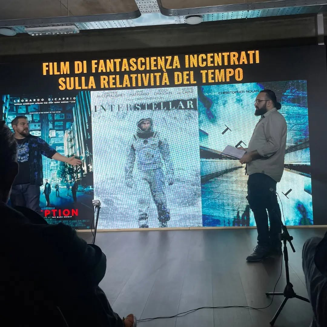 Wearing glasses and holding papers, Vittorio Russo stands beside a screen on which “INTERSTELLAR” is readable.