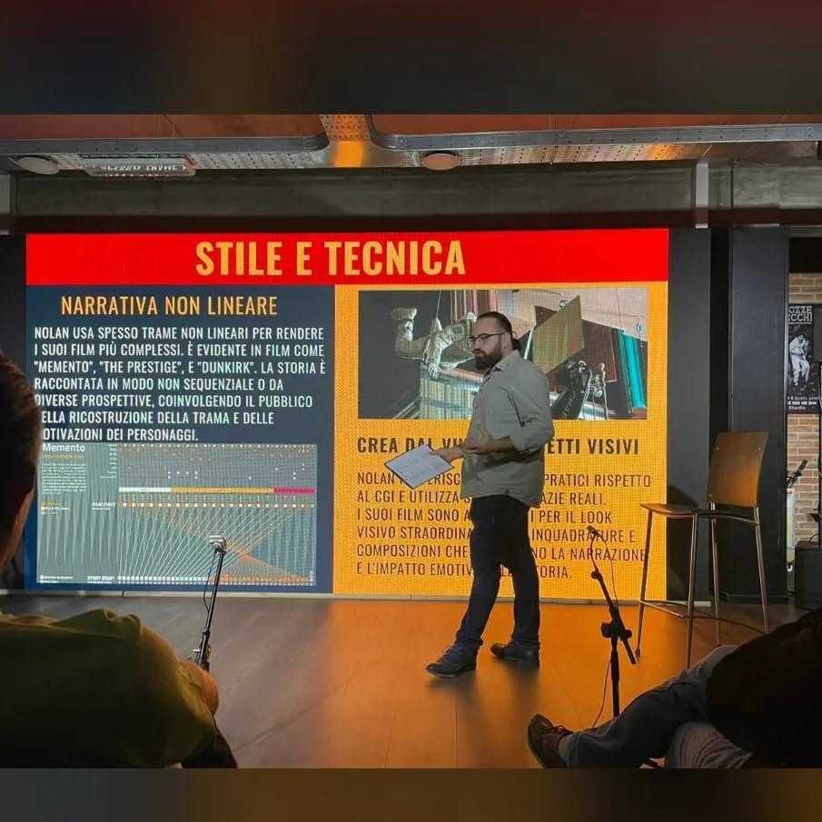 Wearing glasses and holding a sheet, Vittorio Russo speaks in front of the red screen titled “STILE E TECNICA”.