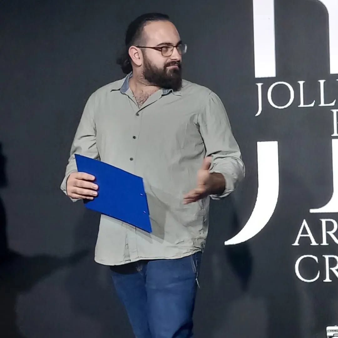 Vittorio Russo, bearded and wearing glasses, holds a blue folder and gestures before a dark backdrop with large white letters, including “JOLL”.