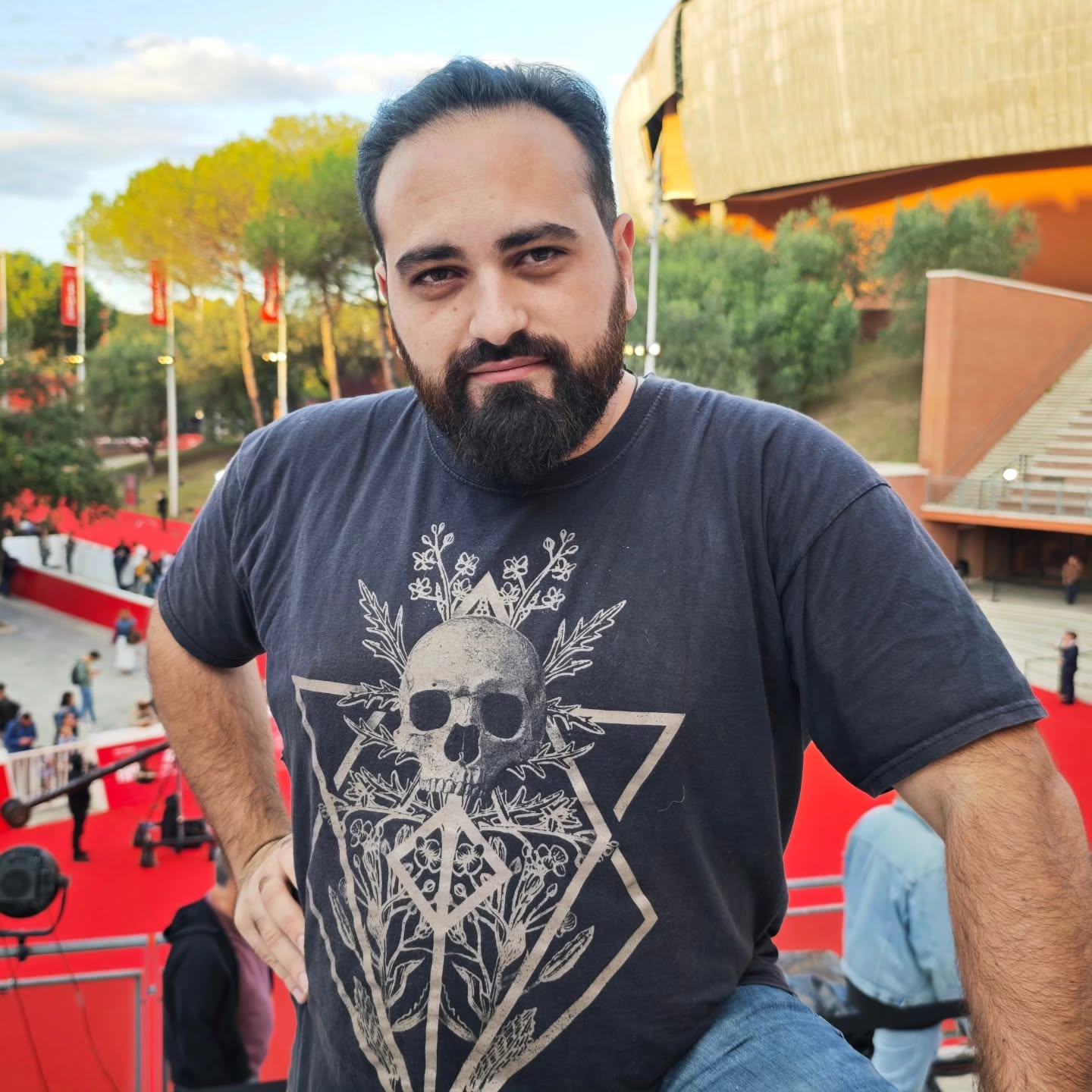 Vittorio Russo, bearded in a dark T-shirt printed with a skull, poses on an outdoor red carpet, with trees and a curved building behind him.