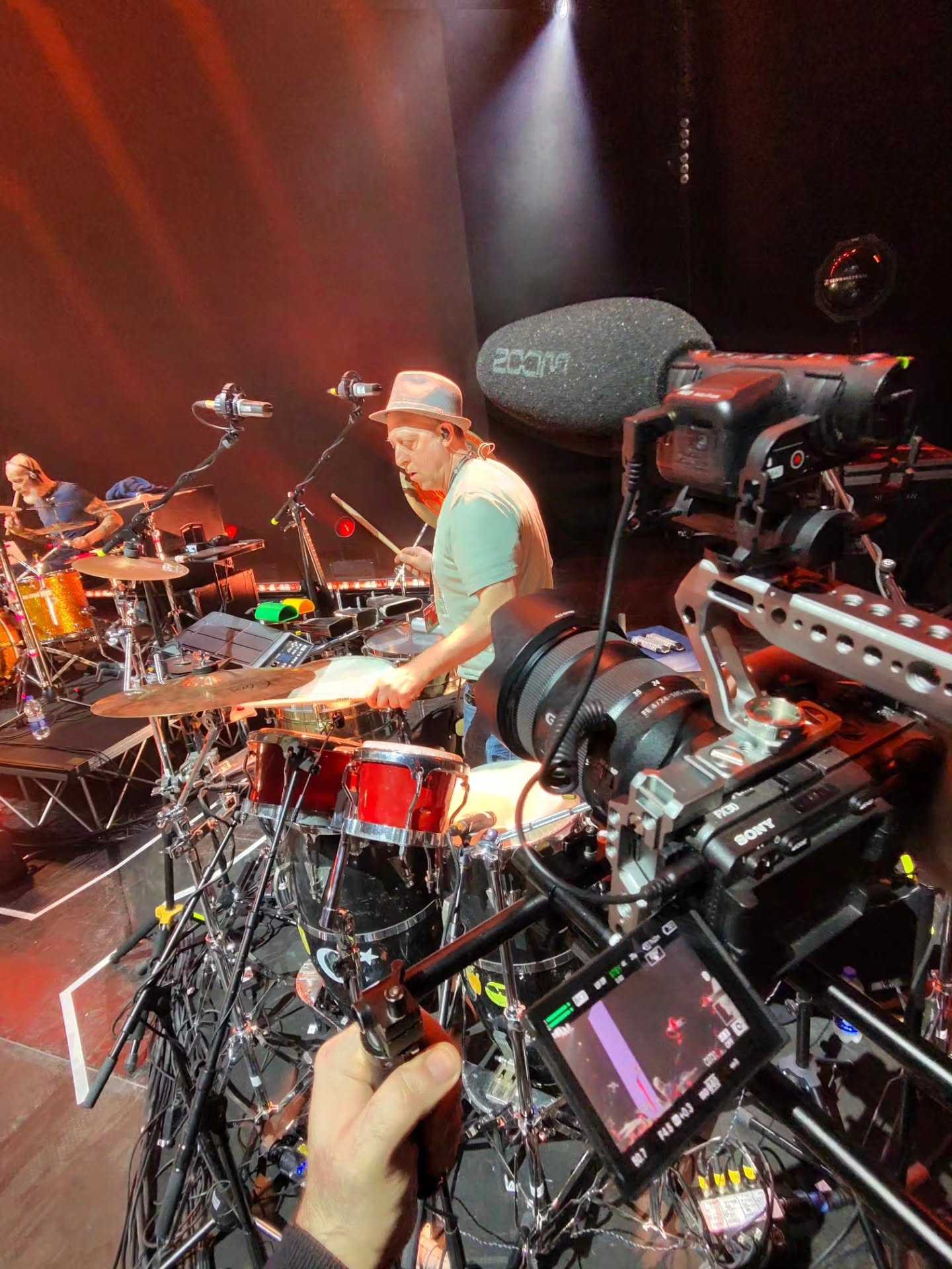 A hand holds a grip beside a cinema camera in the foreground, while a drummer in a hat plays on stage under red lights.