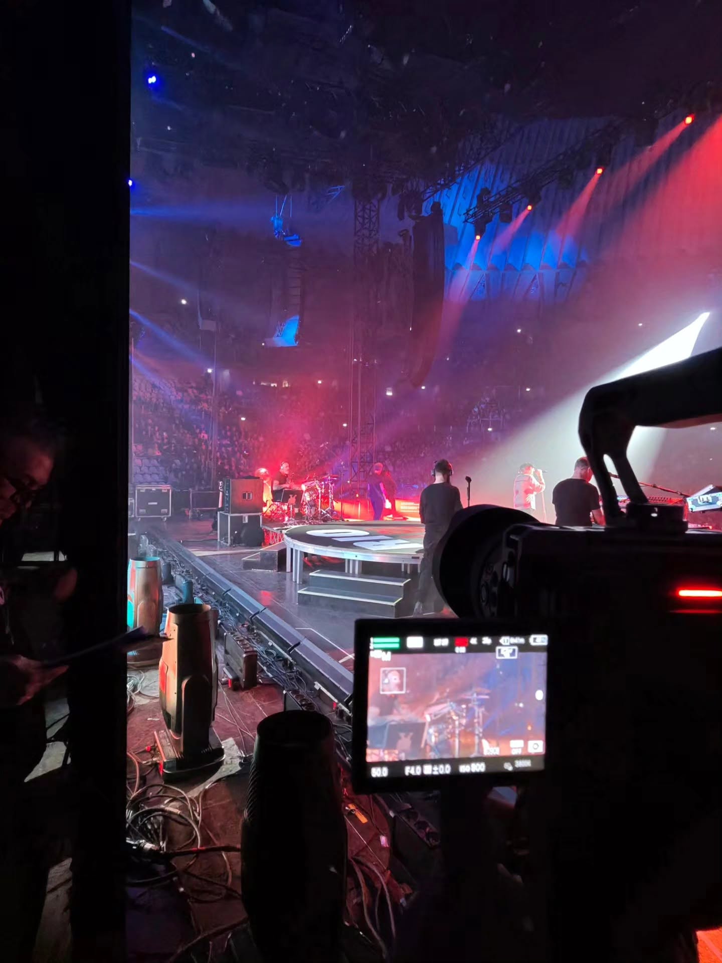 From the edge of the stage, a cinema camera with its monitor on frames a band playing in beams of blue and red light, with the audience in the hall.