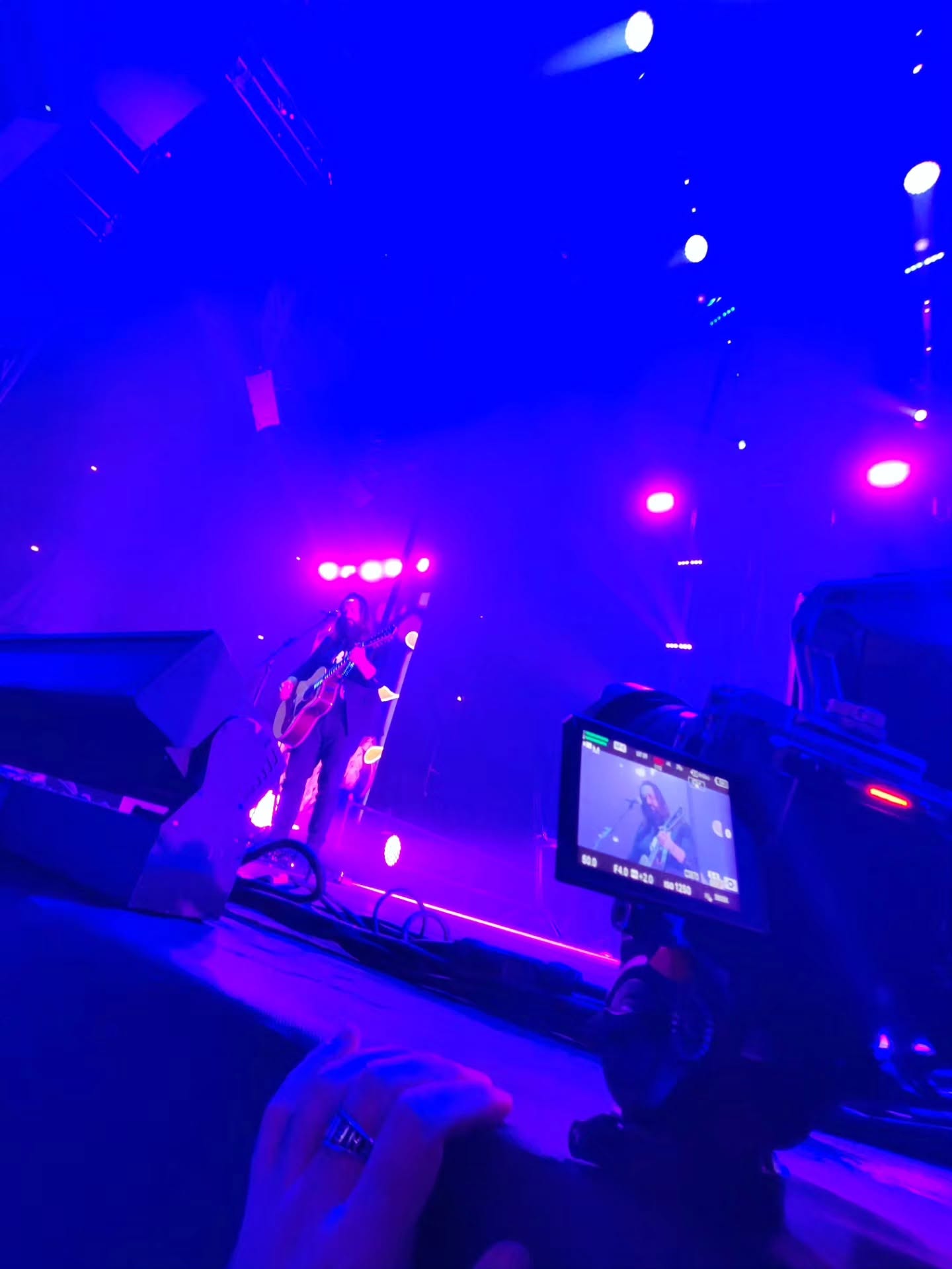 A hand with a ring rests in the foreground near a cinema camera, while a guitarist plays on a stage wrapped in blue and purple light.