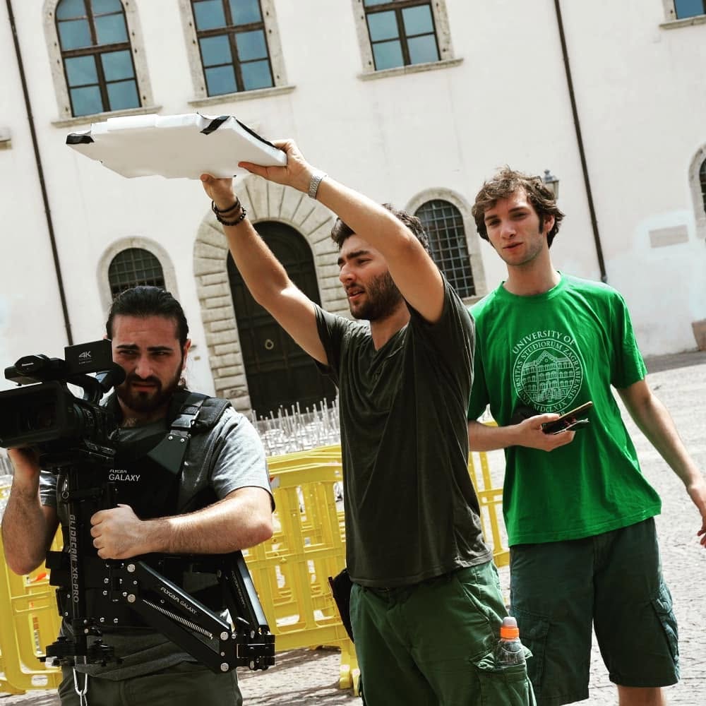Vittorio Russo, bearded, supports a cinema camera on a stabilizer in a courtyard, while one man raises a white board and another stands nearby.
