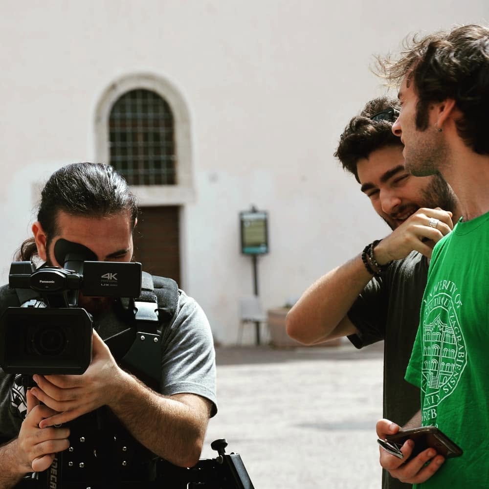 Hands and arms hold a cinema camera to the viewfinder in a courtyard, while two men beside it, one bearded, talk to each other.