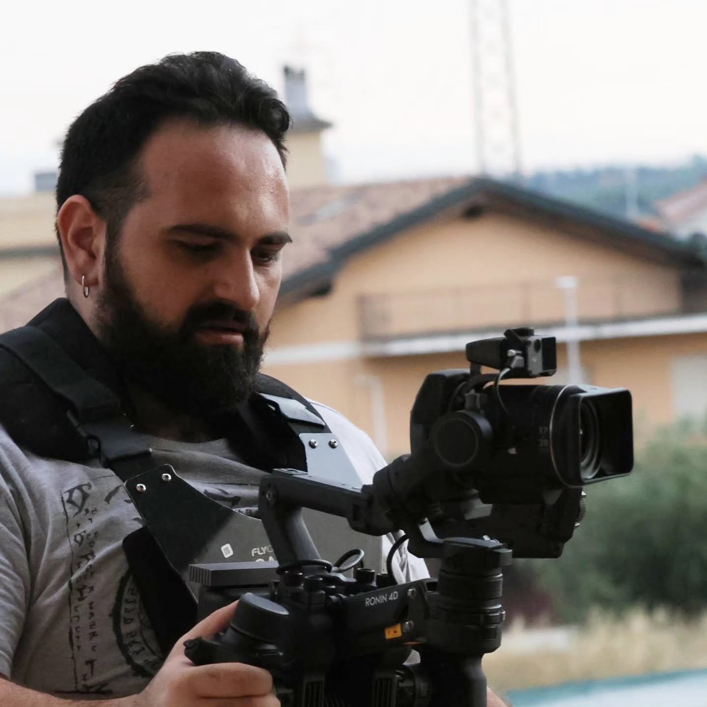 Vittorio Russo, bearded, looks down while holding a cinema camera on a stabilizer, with houses and roofs behind him.