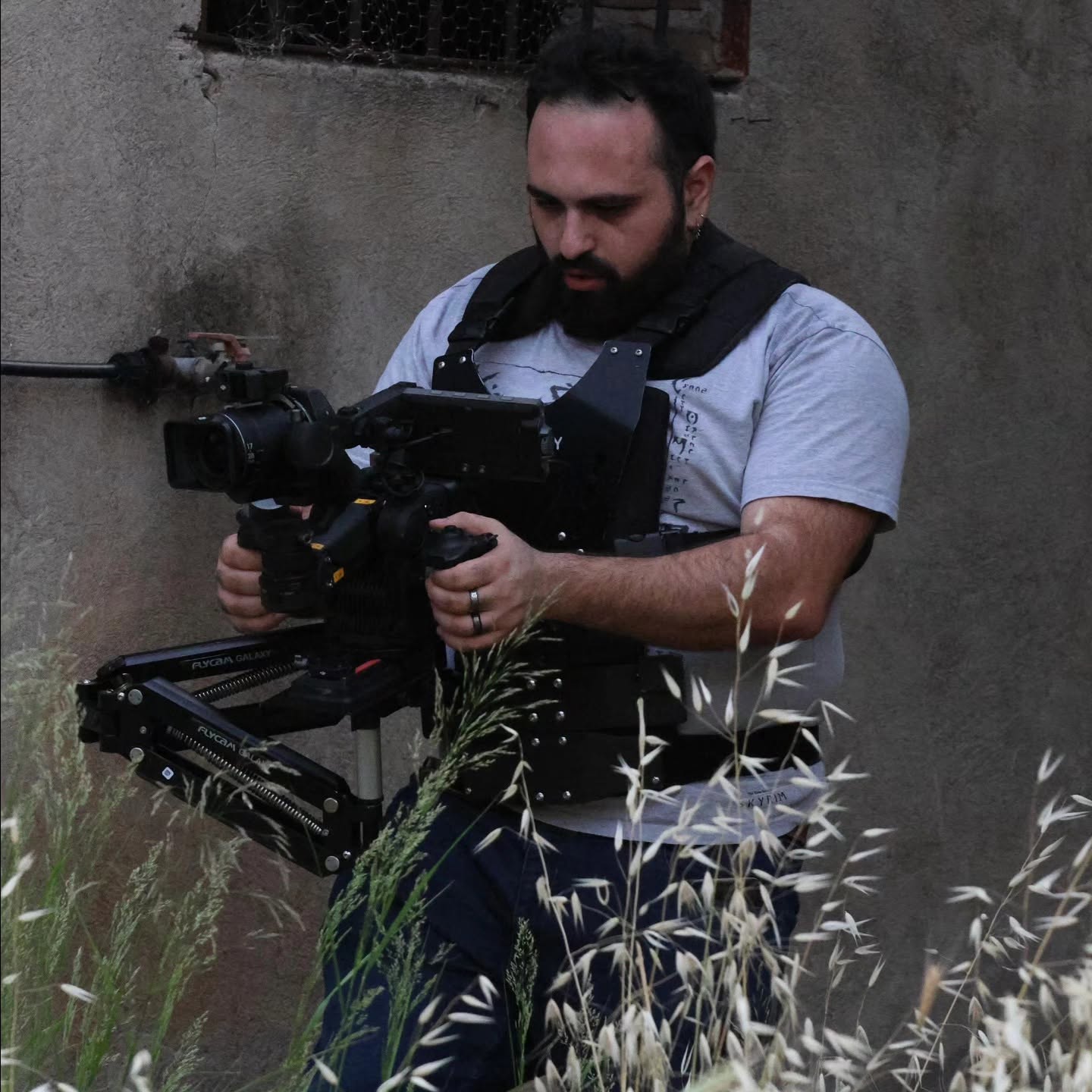 Vittorio Russo, bearded and wearing a support vest, holds a cinema camera on a stabilizer among tall grass, in front of a stone wall.