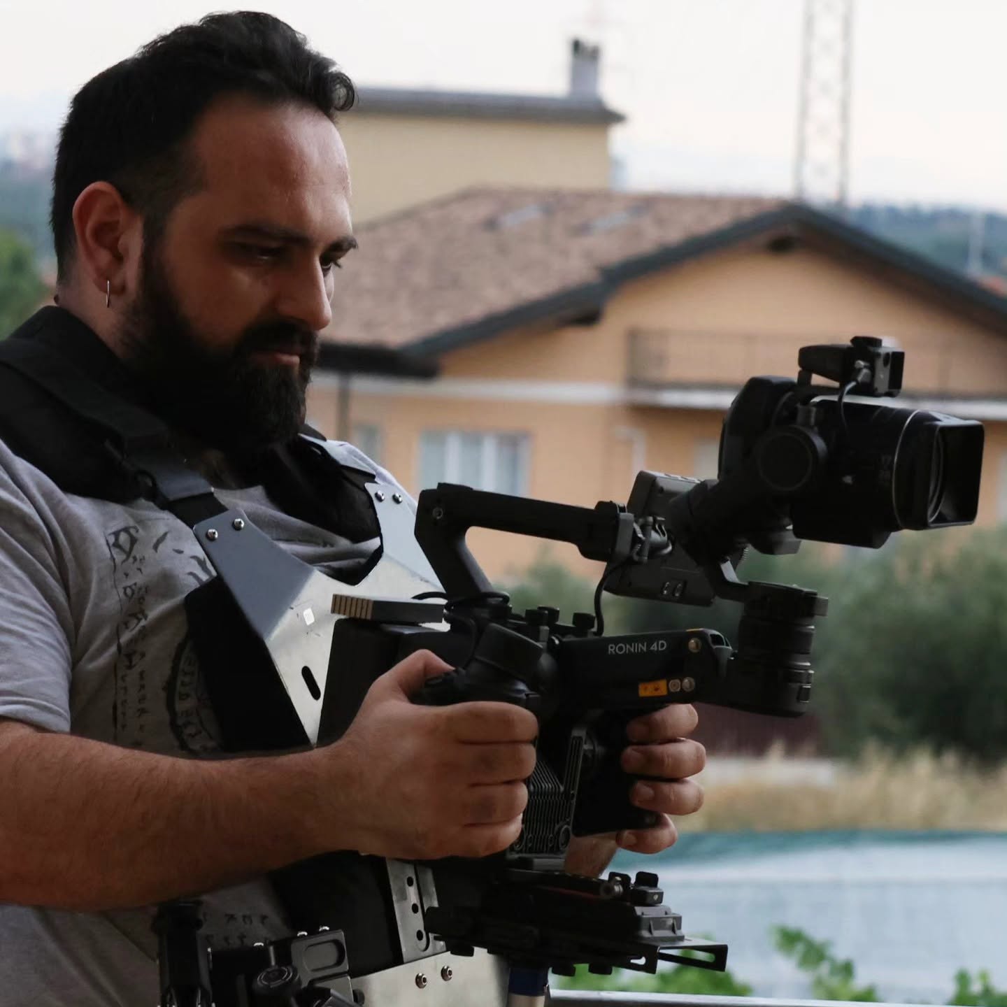 Vittorio Russo, bearded, holds with both hands a cinema camera mounted on a stabilizer, with houses and trees behind him.