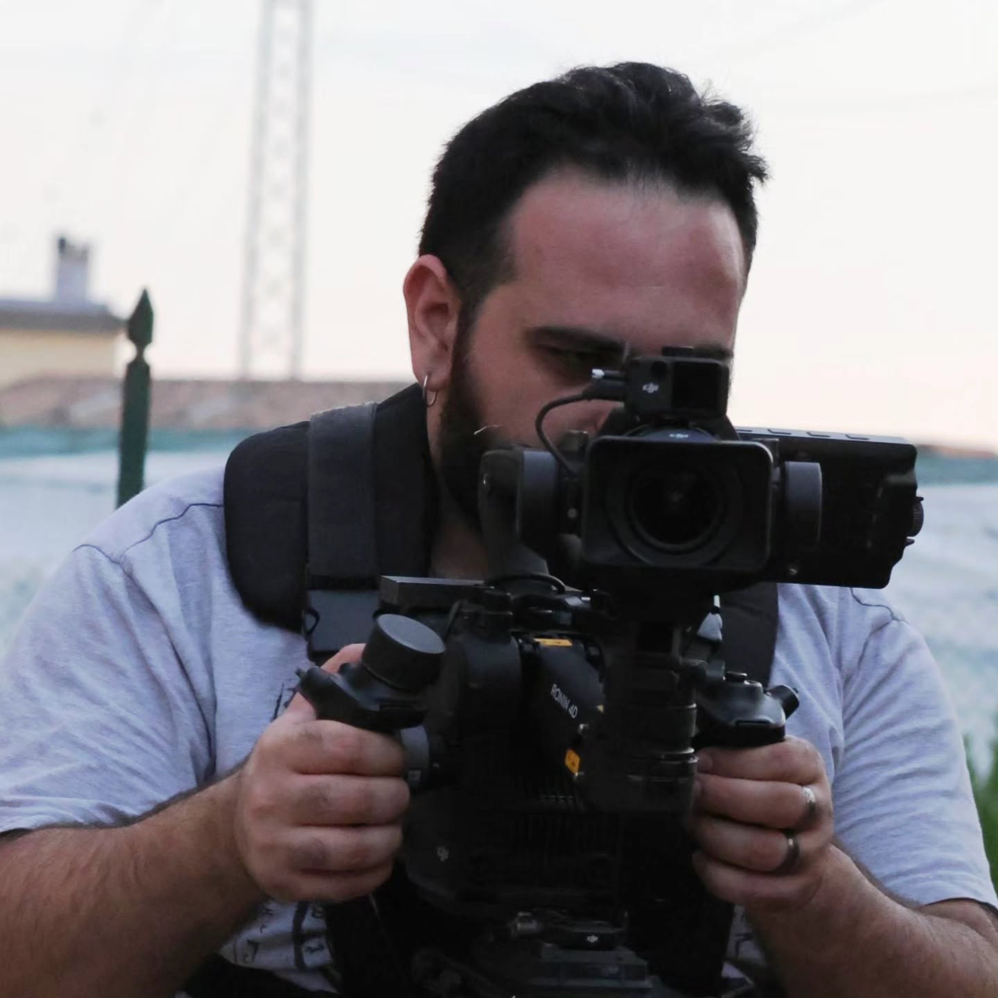 Vittorio Russo, bearded, brings his face close to a cinema camera on a stabilizer and holds it with both hands outdoors.