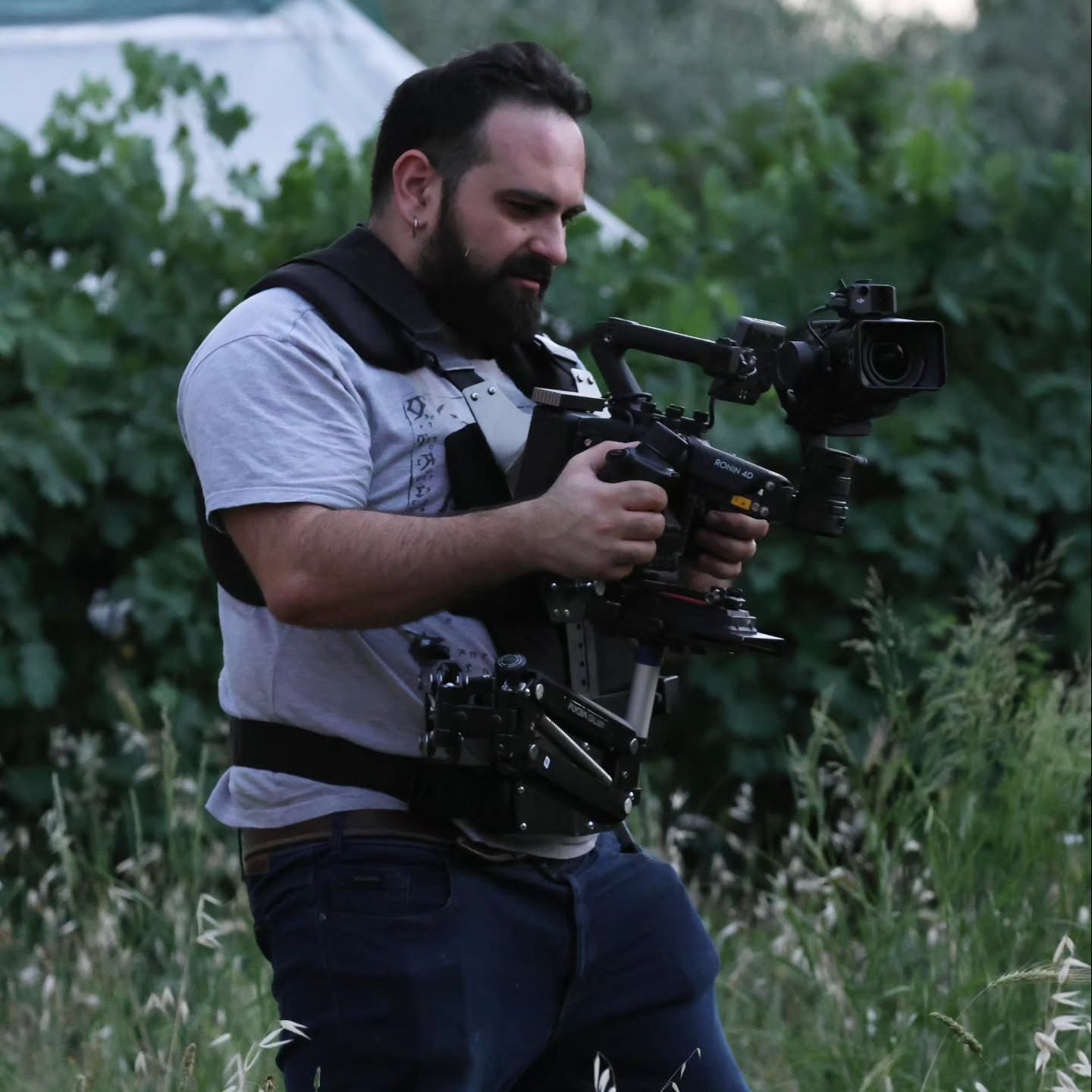 Vittorio Russo, bearded and wearing a support vest, walks through the grass with a cinema camera on a stabilizer, surrounded by green leaves.