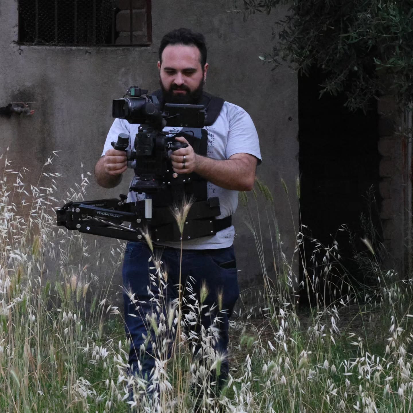 Vittorio Russo, bearded, stands among tall dry grass and adjusts a cinema camera on a stabilizer in front of a stone building.