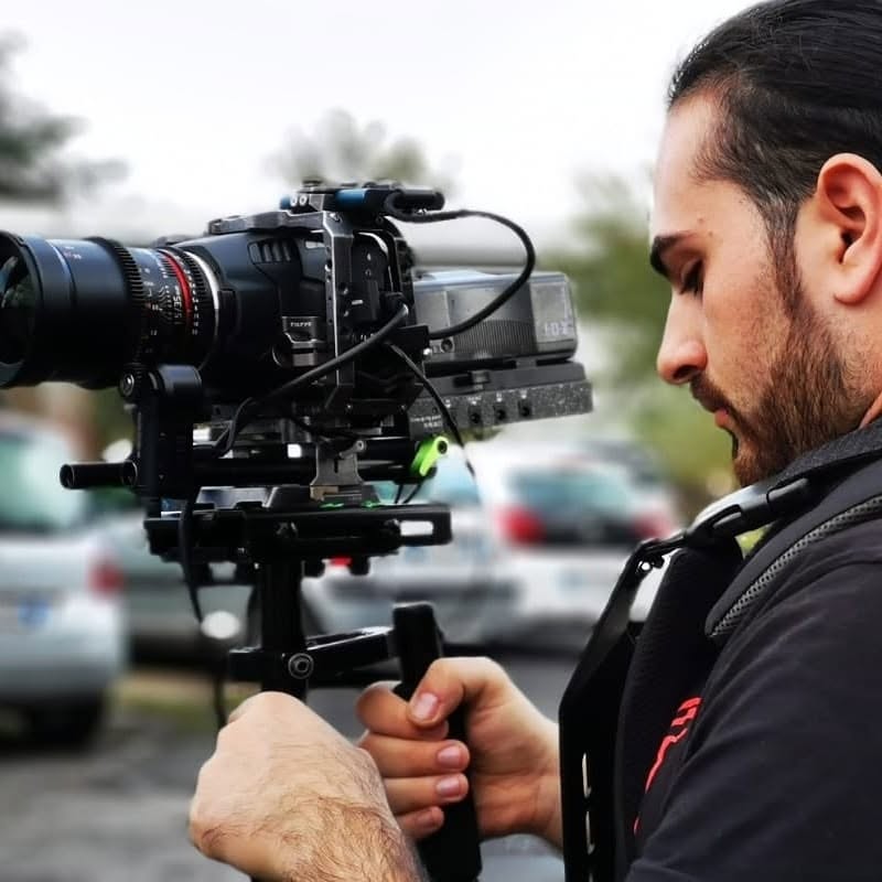 In profile, Vittorio Russo, bearded, grips the handle of a cinema camera on a stabilizer, with cars and trees blurred behind him.