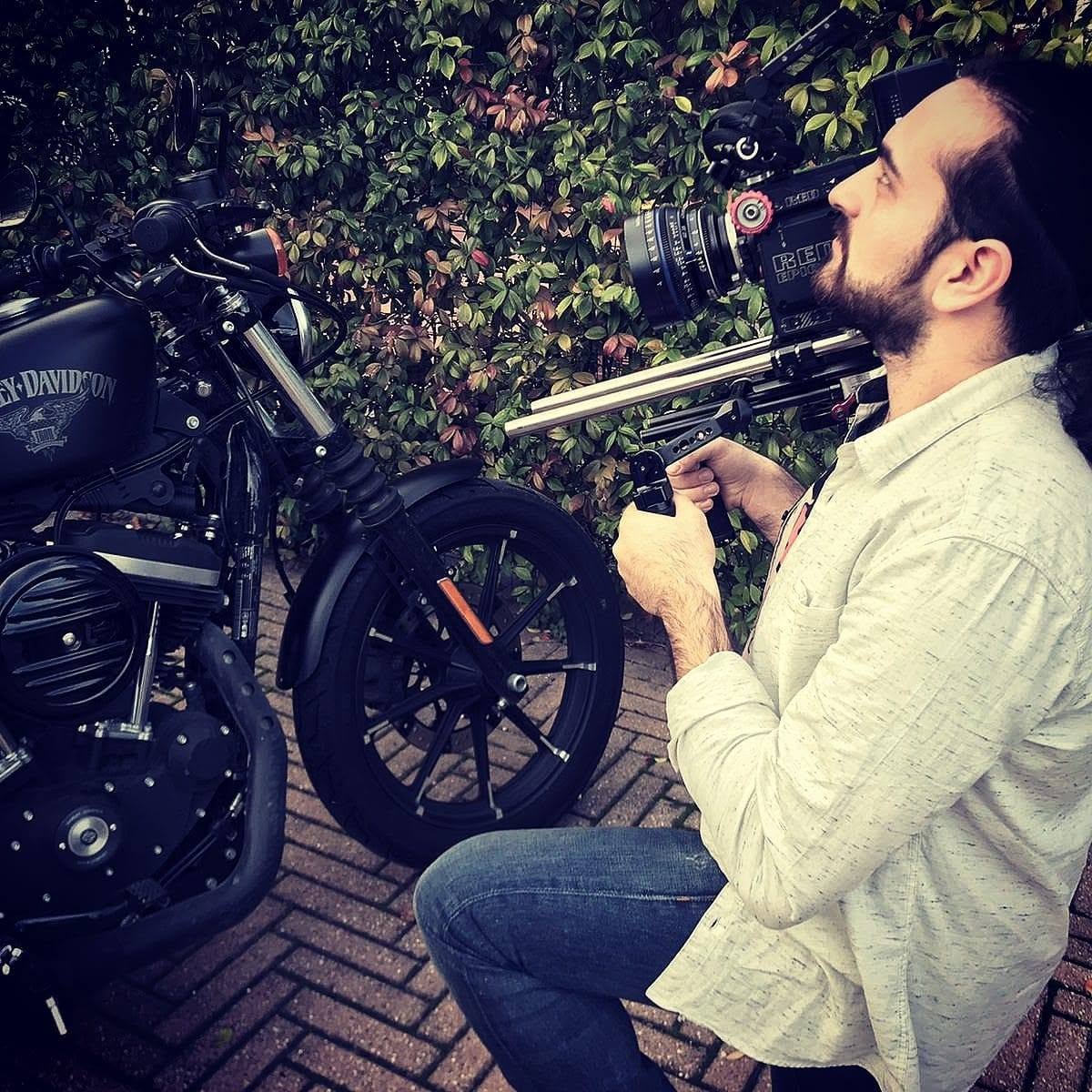 Vittorio Russo, bearded, crouches beside a black motorcycle and holds a cinema camera on a rig, in front of a hedge.