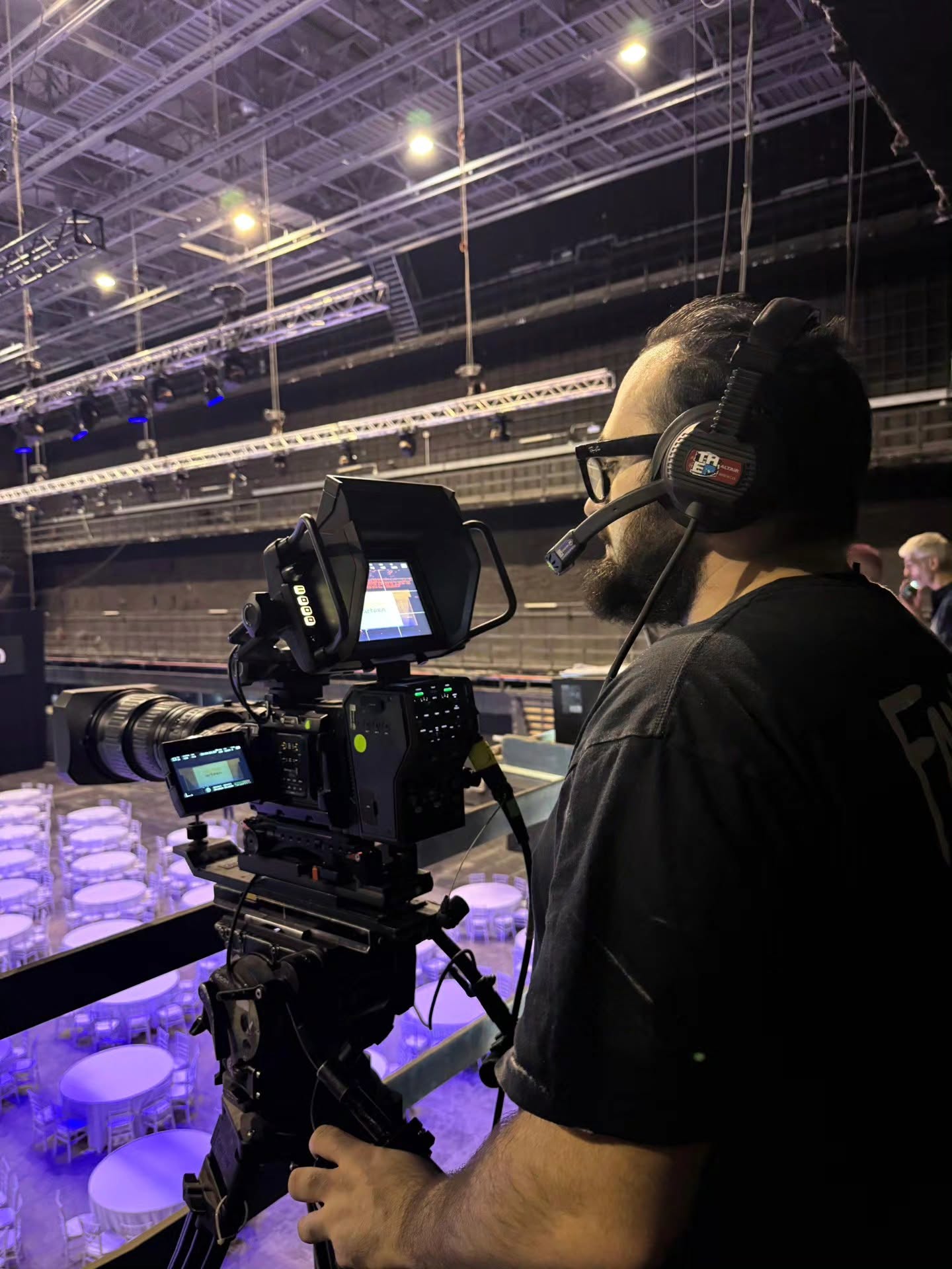 In profile, Vittorio Russo, with a beard, glasses and a headset, stands beside a cinema camera on a tripod aimed at a hall of round tables.