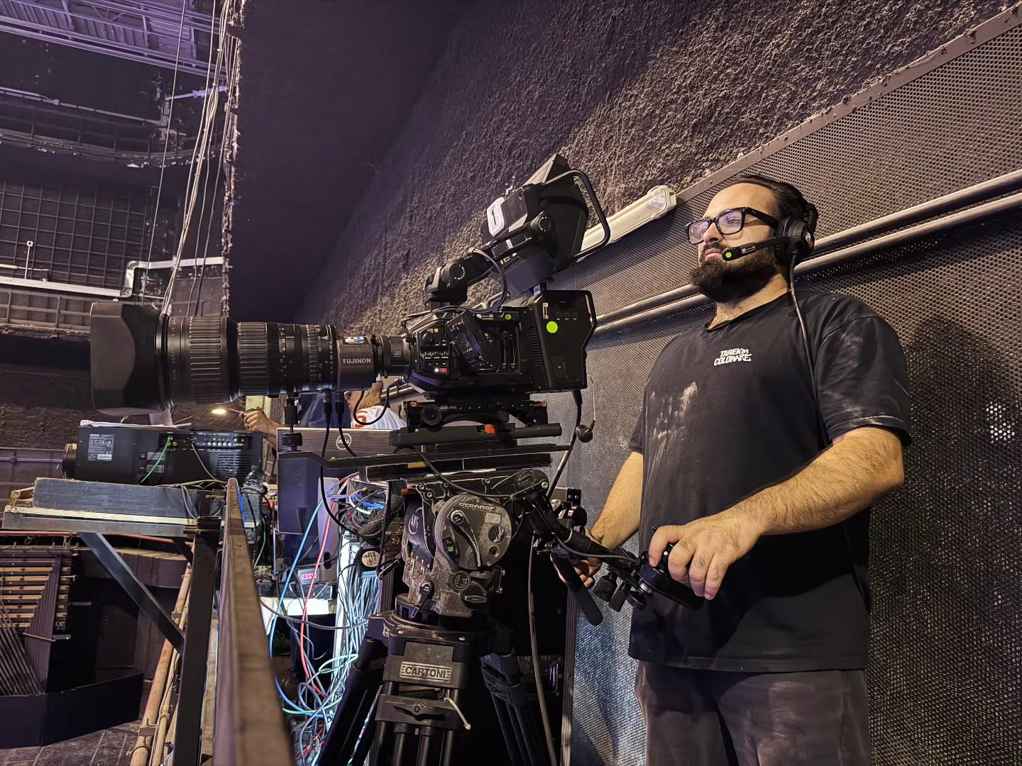 Vittorio Russo, with a beard, glasses and a headset, holds a controller beside a large cinema camera on a tripod in a technical space.
