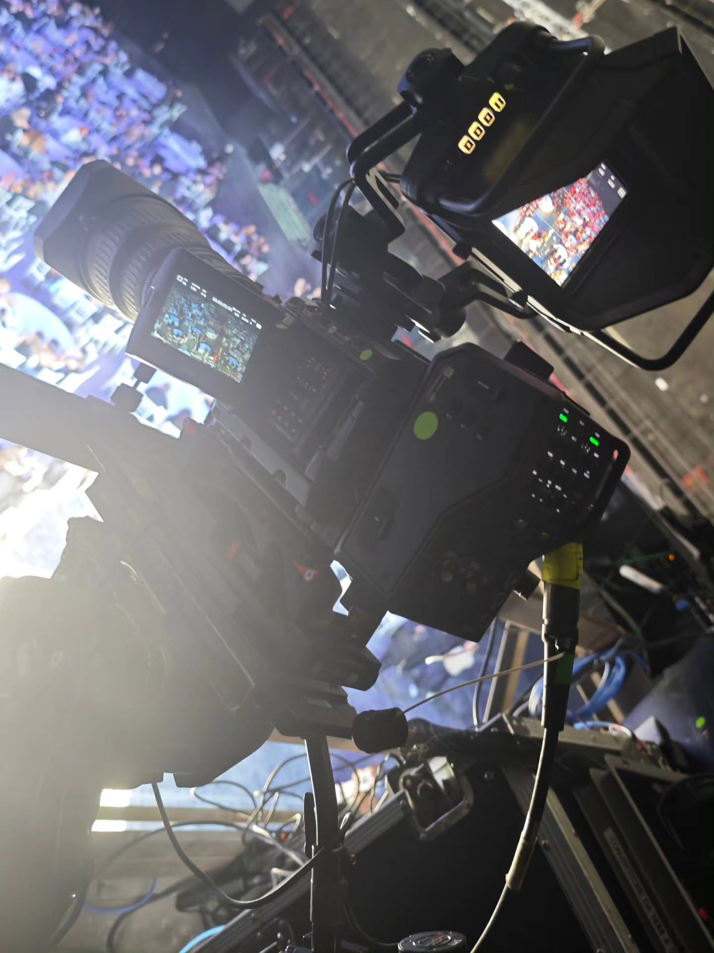 Against the light, a broadcast camera with monitors and cables frames a lit hall filled with an audience.